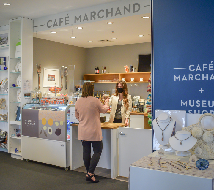 One visitor buying a beverage from an employee at Café Marchand, located in the Westmoreland Museum Shop. In the foreground to the right are several necklaces, bracelets, earrings, and a handbag for sale at the Museum Shop.