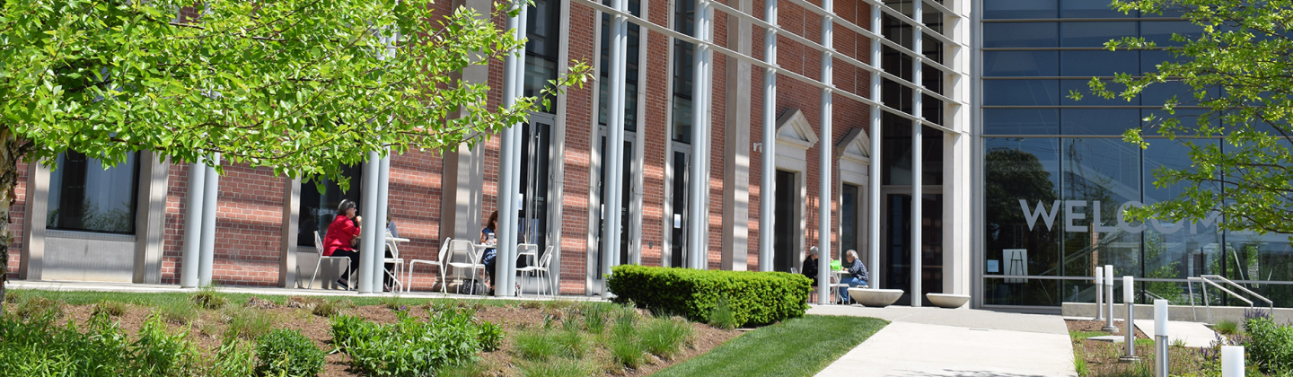 Visitors sit at white tables positioned near the Museum's brick walls and entry doors on the terrace during a bright sunny day.