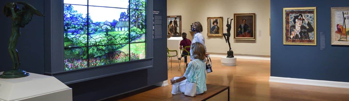 Two visitors, one sitting on a bench and one standing, observing Tiffany’s Thomas Lynch Window in The Westmoreland’s 20th Century gallery. In the background are other portraits and sculptures in the gallery.