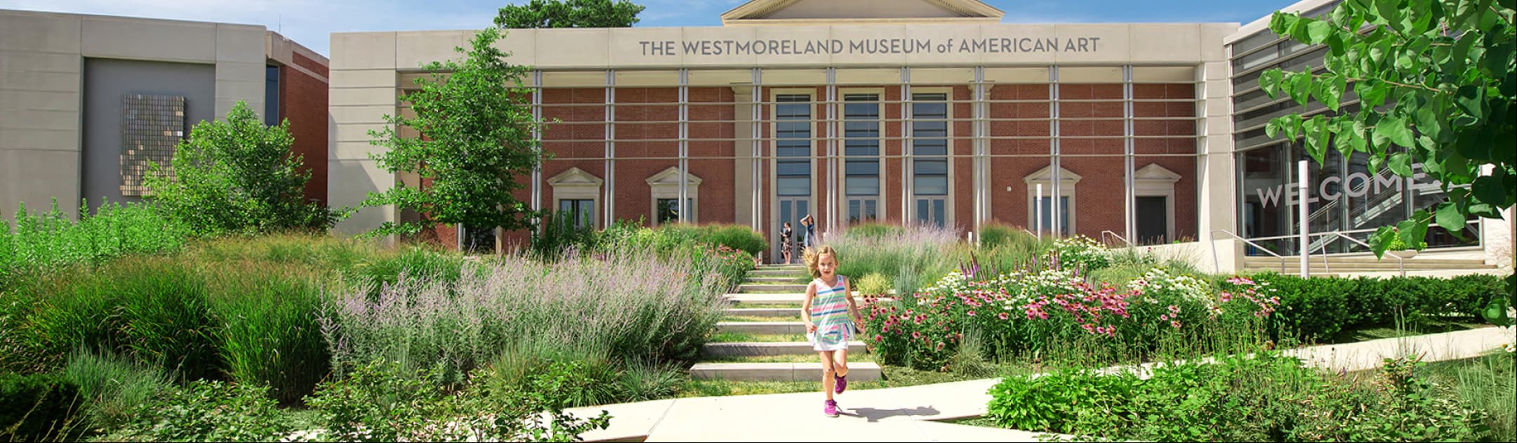 A young girl running down The Westmoreland Museum of American Art’s concrete front steps towards the camera, with The Museum’s front entrance in the background, surrounded by bushes, trees, and flowers.
