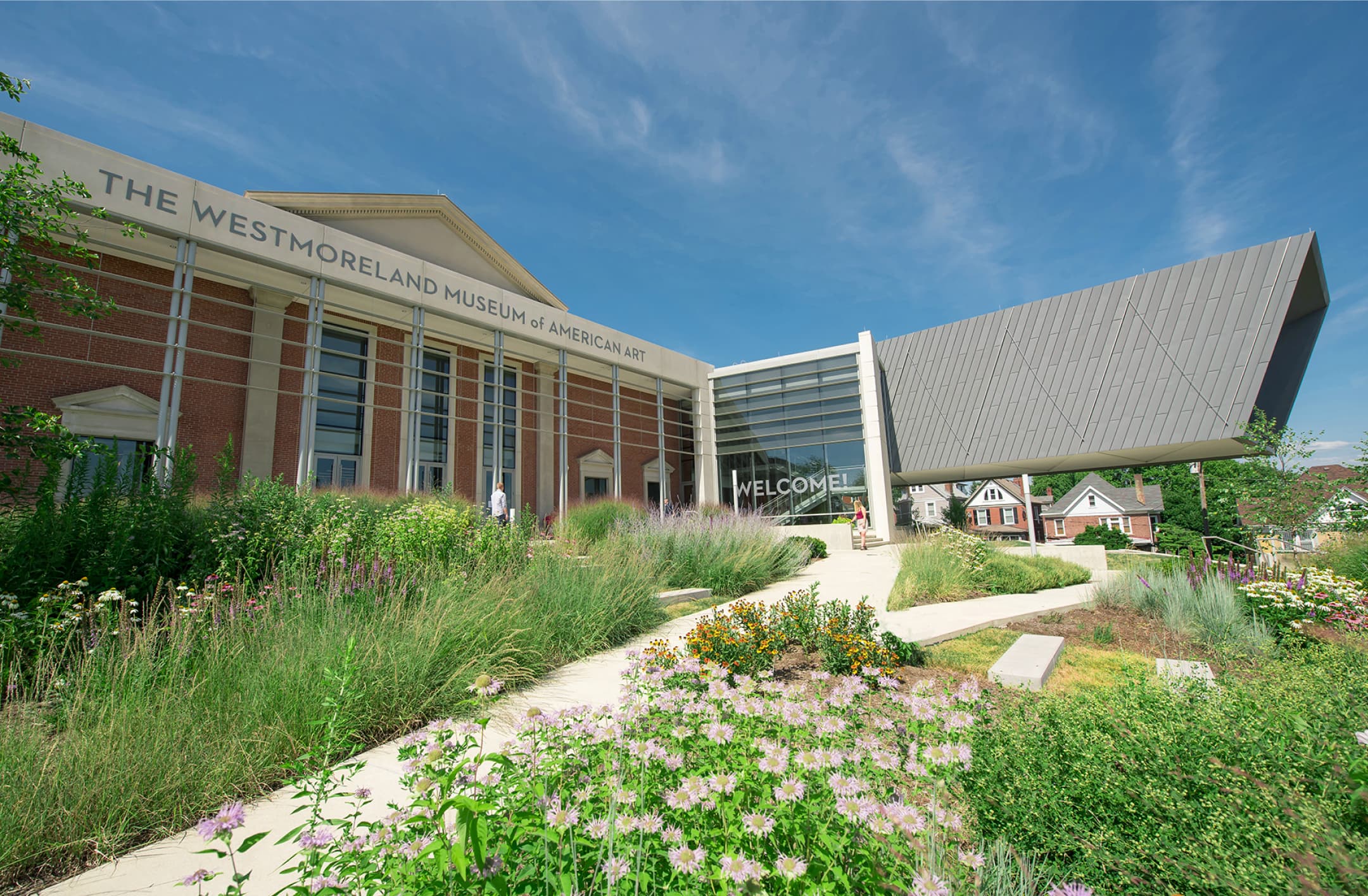 To the left, the exterior of the Westmoreland Museum of American Art’s front entrance. To the right, the exterior of the museum’s Cantilever Gallery. In the foreground are bushes, flowers, and a pathway to the Museum’s front entrance.