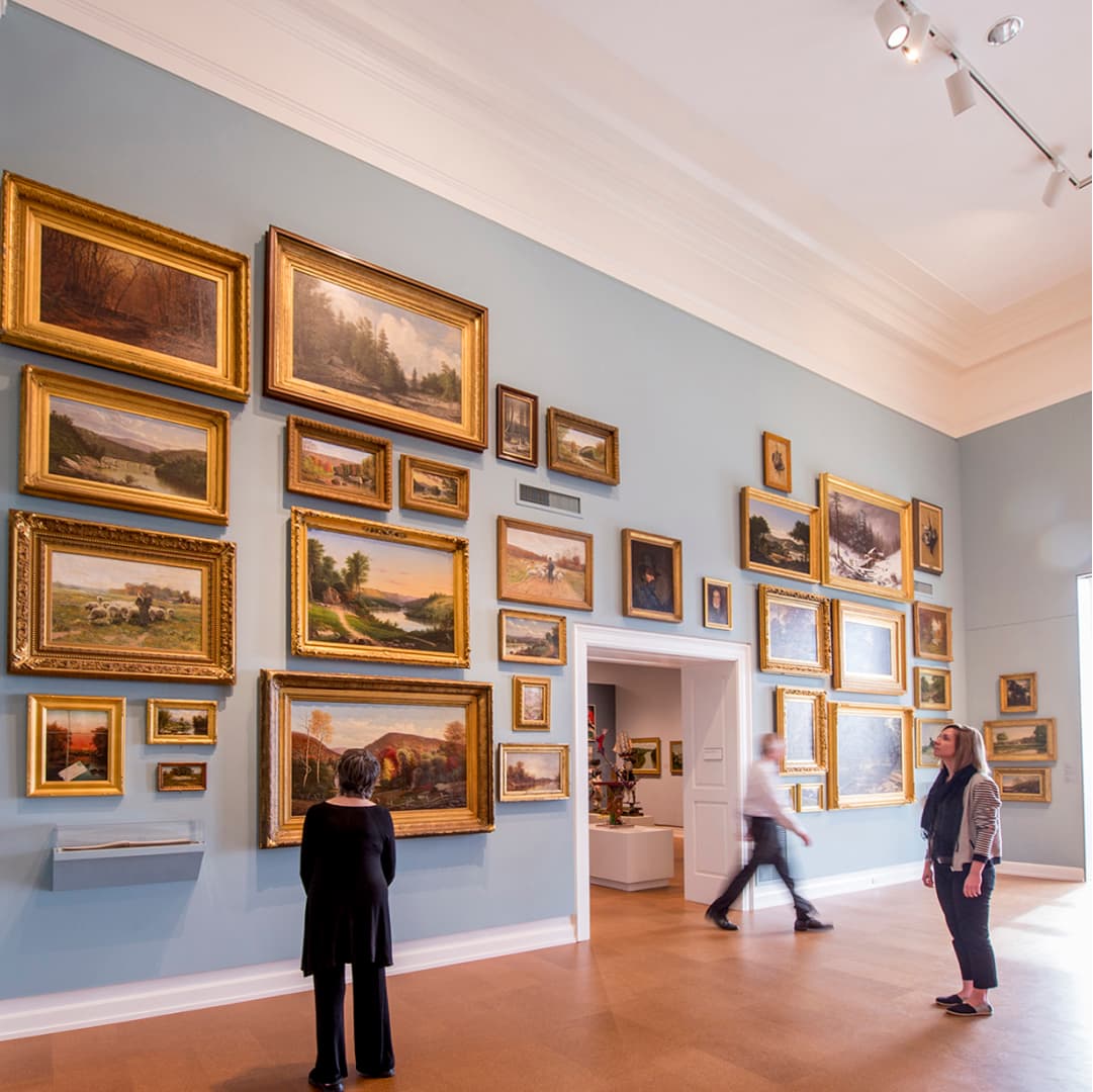 Three visitors, two standing and one walking, observing the 18th and 19th century paintings of Pennsylvania landscapes in varying sizes and shapes hung salon style in The Westmoreland’s McKenna gallery.