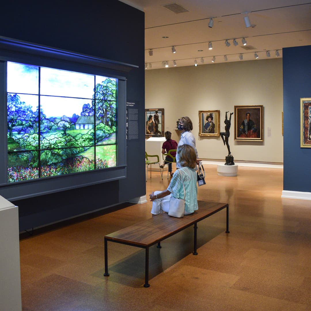 Two visitors, one sitting on a bench and one standing, observing Tiffany’s Thomas Lynch Window in The Westmoreland’s 20th Century gallery. In the background are other portraits and sculptures in the gallery.